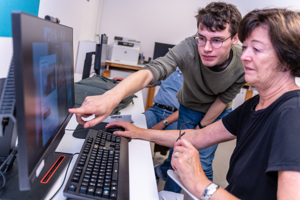 Ein junger Mann mit Brille zeigt einer älteren Frau etwas auf einem Computerbildschirm. Sie sitzt an einem Schreibtisch, hält eine Brille in der Hand und tippt auf der Tastatur, während er sich über sie beugt und auf den Monitor deutet.